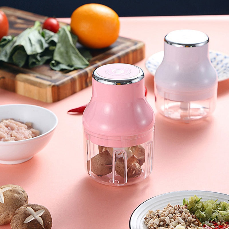 A Pink and a White Mini Electric Food Chopper surrounded by other ingredients and utensils. The pink one has some ingredients inside ready to be chopped